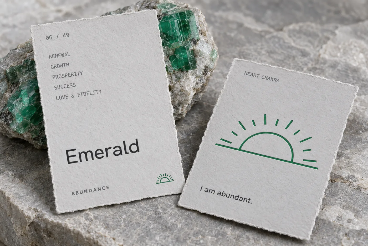 Concept photograph of two torn-edge paper stone cards beside raw emerald: the front reads 06 of 49, Emerald, Abundance, and the back carries a green sunrise and the line I am abundant
