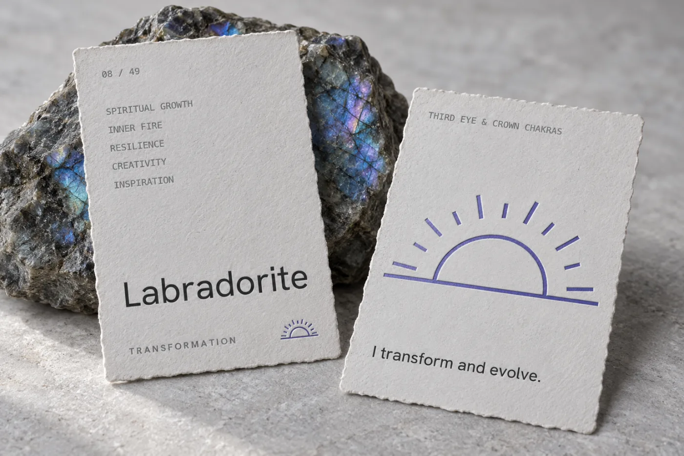 Concept photograph of two torn-edge paper stone cards beside raw labradorite: the front reads 08 of 49, Labradorite, Transformation, and the back carries a violet sunrise and the line I transform and evolve