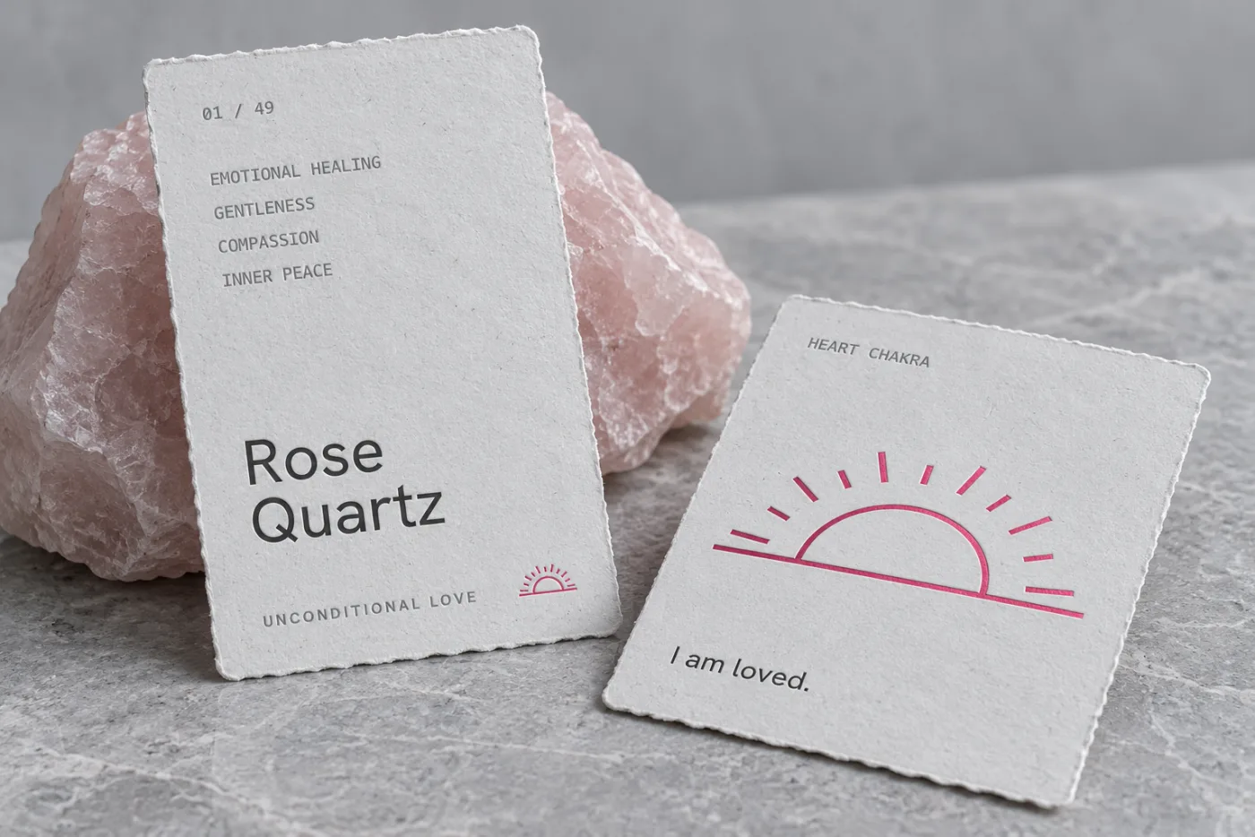 Concept photograph of two torn-edge paper stone cards beside raw rose quartz: the front reads 01 of 49, Rose Quartz, Unconditional Love, and the back carries a pink sunrise and the line I am loved